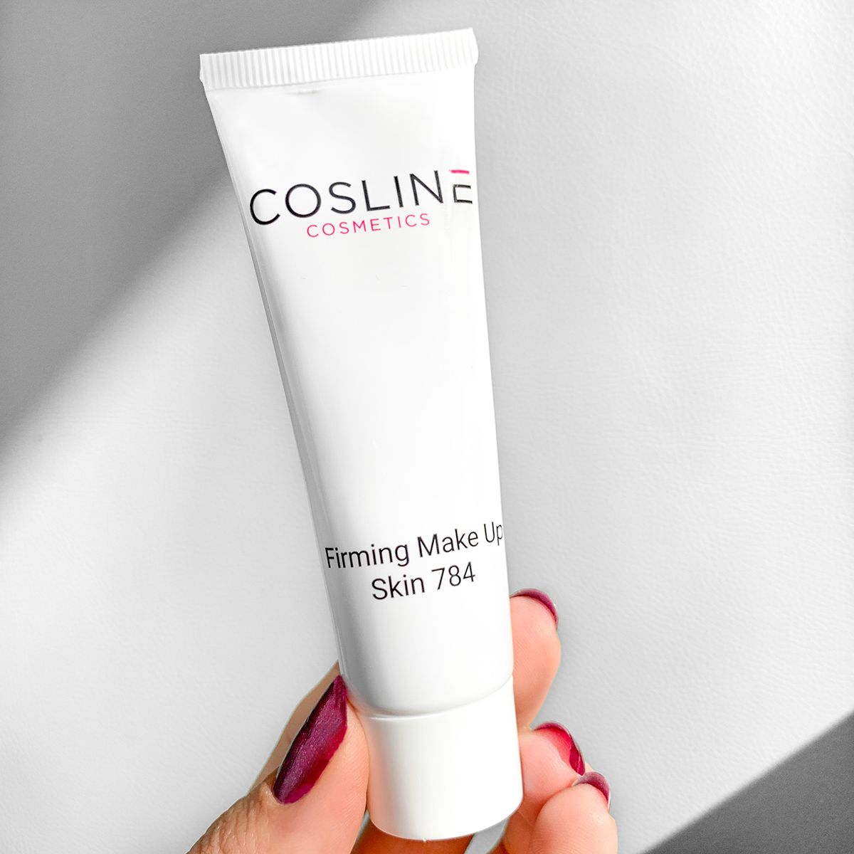 Eine Hand hält eine weiße Tube Cosline Firming Make Up, Skin 784, mit rotem Nagellack.