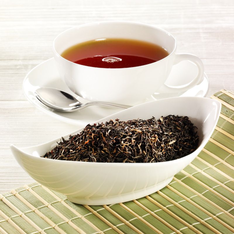Weiße Tasse mit Tee, Untertasse, Löffel und Schale mit losem Tee auf Bambusmatte.