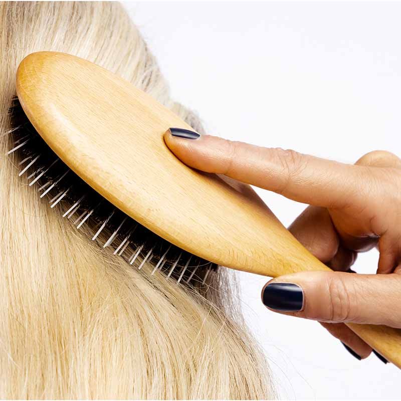 Hand streicht mit ovaler Haarbürste aus Holz mit Wildschweinborsten durch blondes Haar.