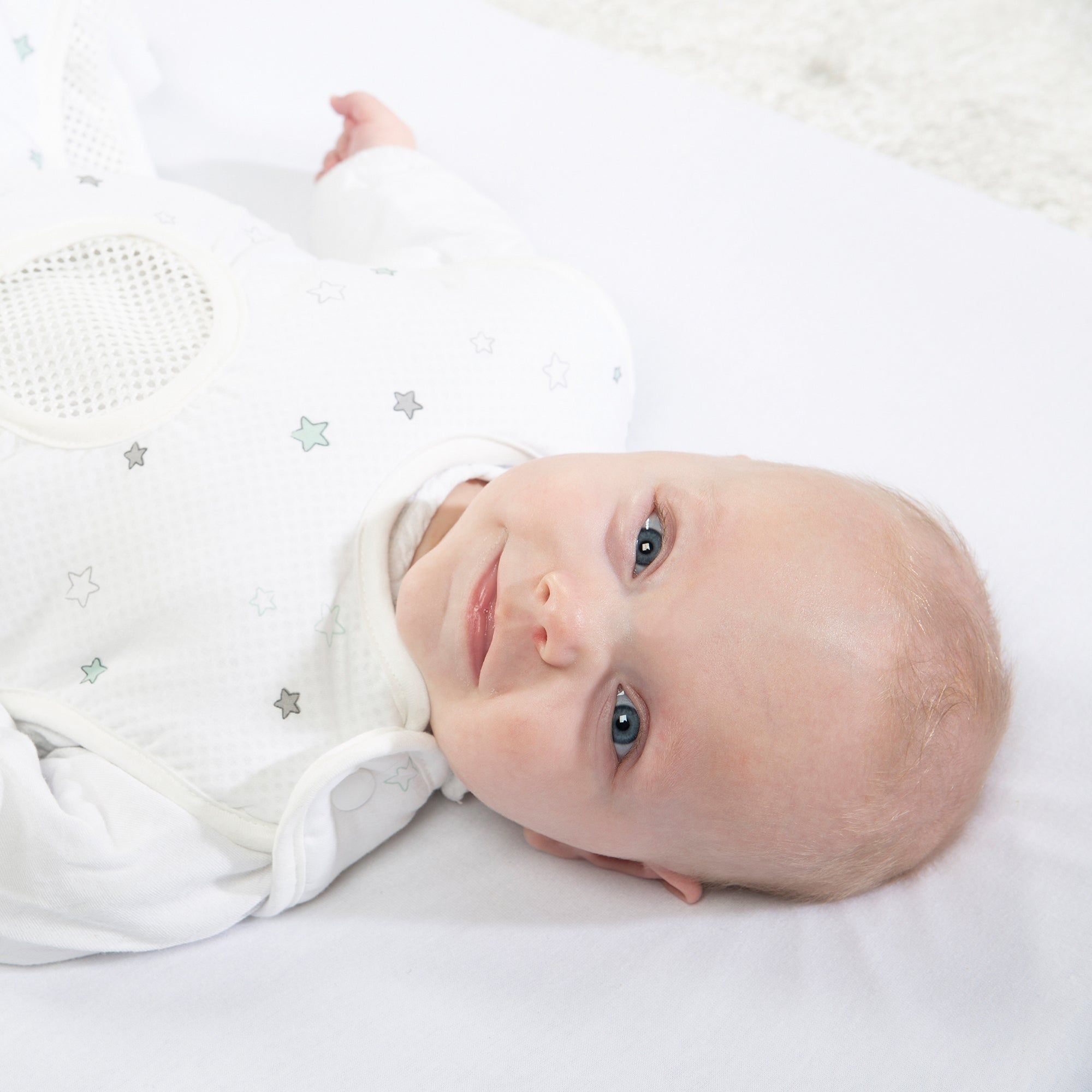 Baby liegt in weißem Schlafsack mit Sternenmuster. Mesh-Einsatz im Brustbereich. Baby schaut in die Kamera.