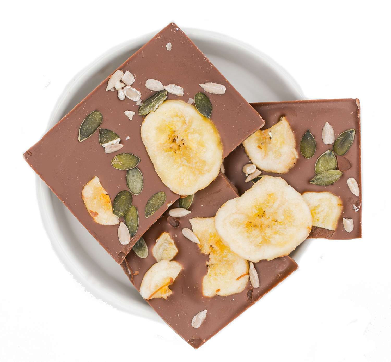 Schokoladenstücke mit Bananenscheiben, Kürbiskernen und Sonnenblumenkernen in einer weißen Schale. Draufsicht.
