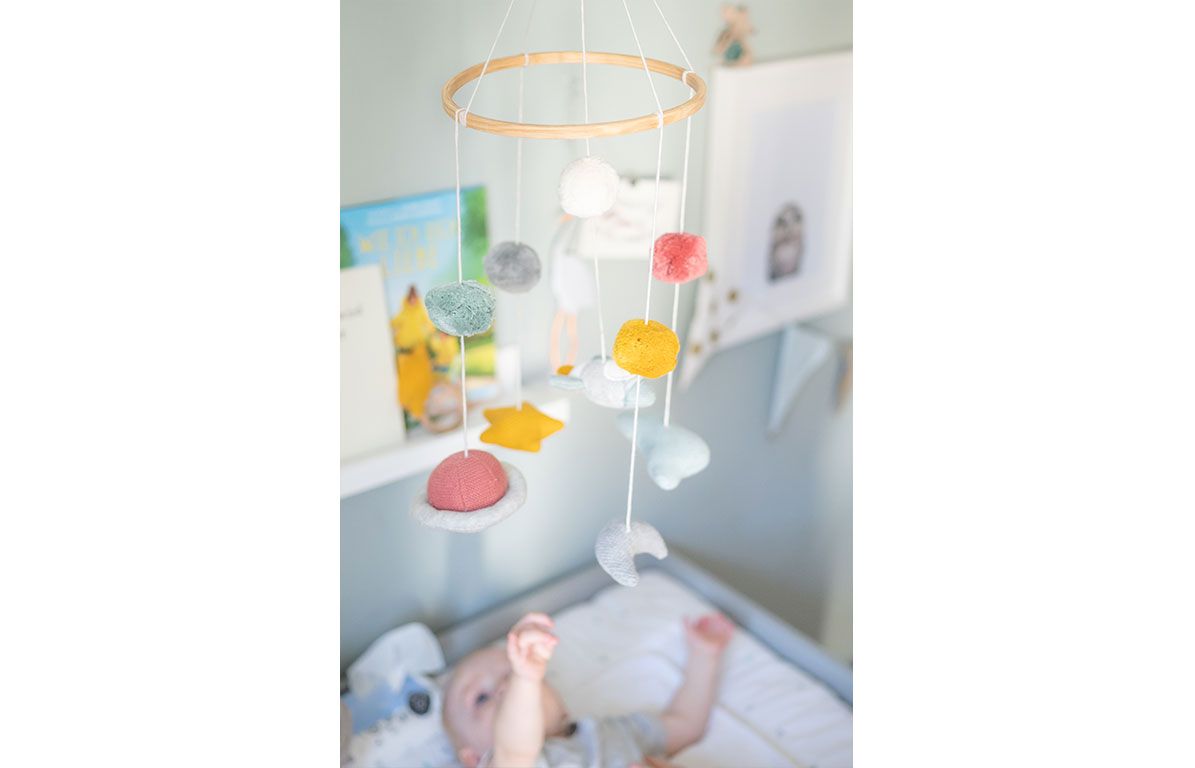 Mobile über Babybett. Holzring mit bunten Kugeln, Sternen und Planeten. Baby liegt auf Wickelauflage.