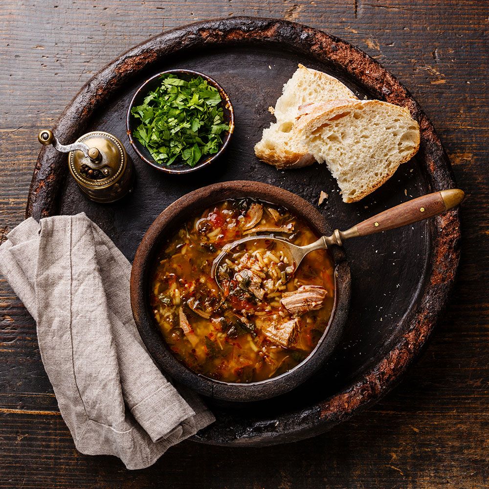 Suppe in einer Schüssel. Brot, Kräuter und Gewürze daneben. Auf einem Holztablett. Dunkler Hintergrund.