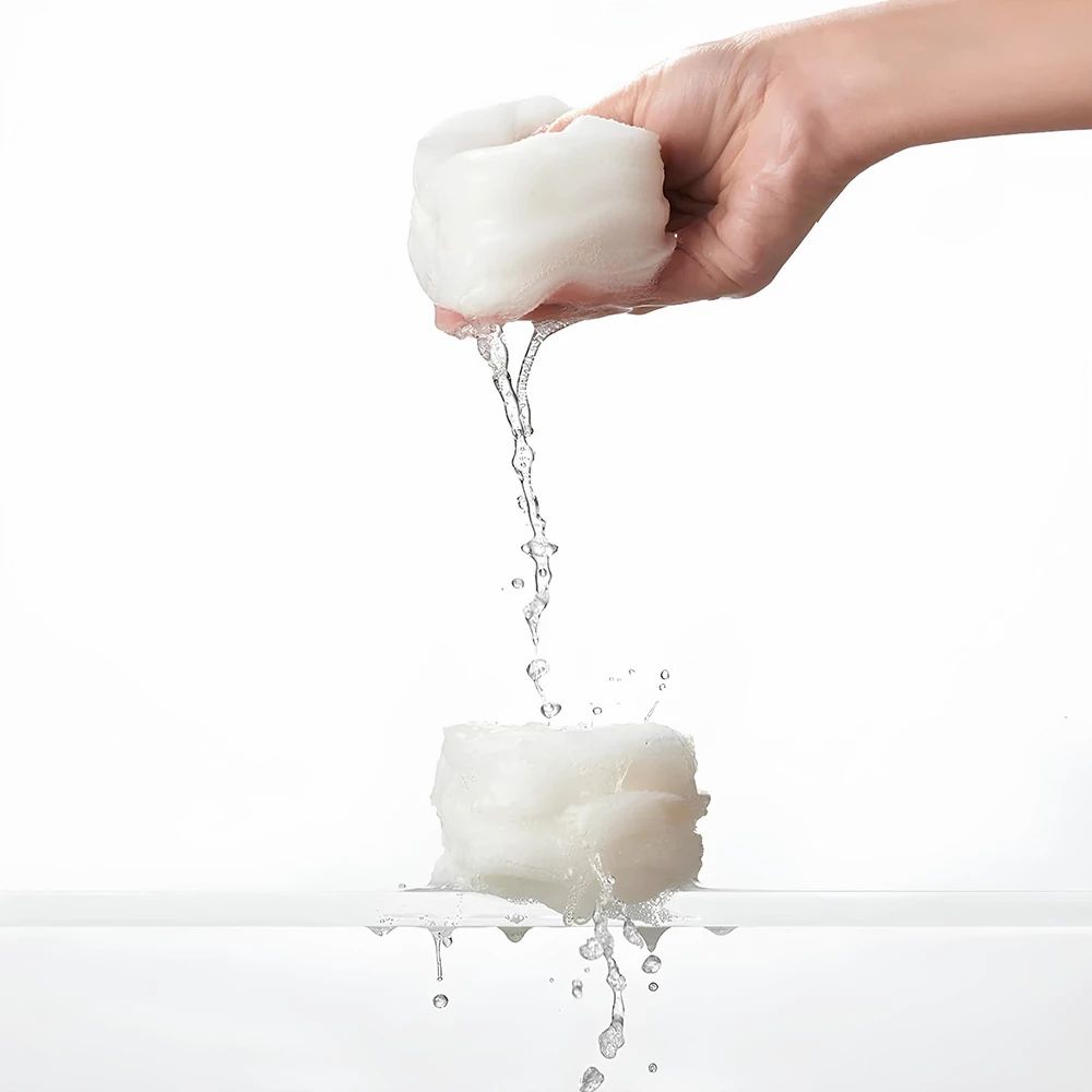 Hand hält ein Pad, Wasser fließt heraus. Pad ist weiß und rund. Wasser spritzt. Hintergrund weiß.