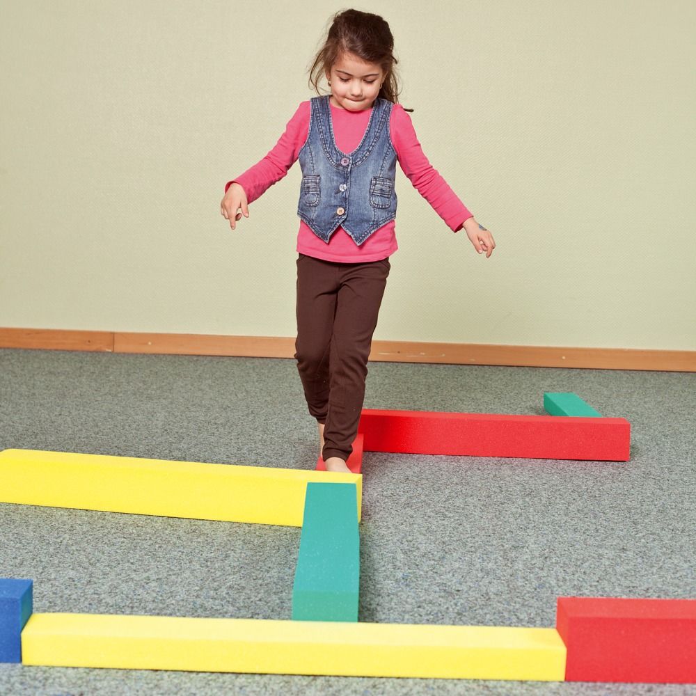 Kind balanciert auf gelben, grünen und roten Schaumstoffbausteinen. Weitere farbige Bausteine liegen auf dem Boden.