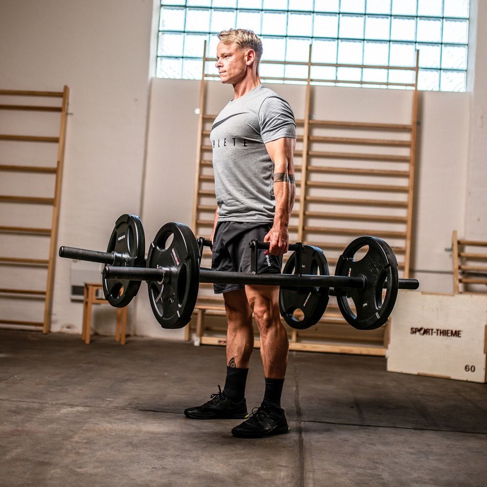 Mann mit Farmers Walk Bar mit Gewichten. Schwarze Stange mit Griffen und Gewichtsscheiben. Sport-Thieme Logo auf Kiste.