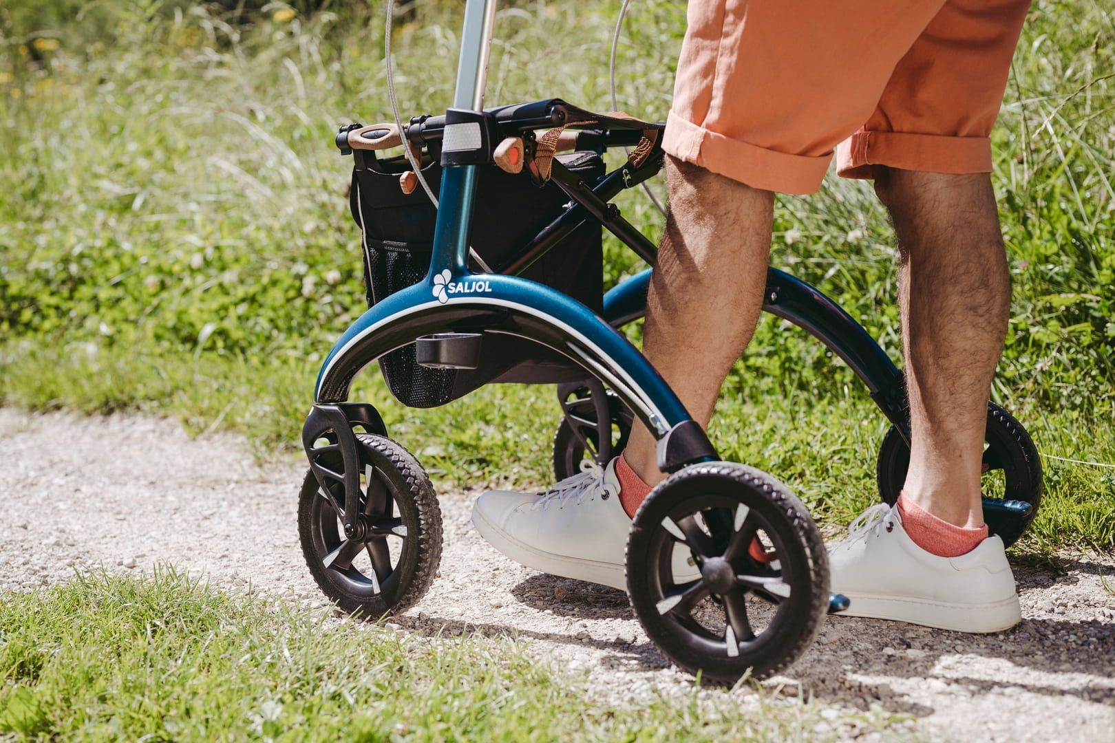 Person steht neben einem blauen Rollator auf einem Weg. Der Rollator hat vier Räder.