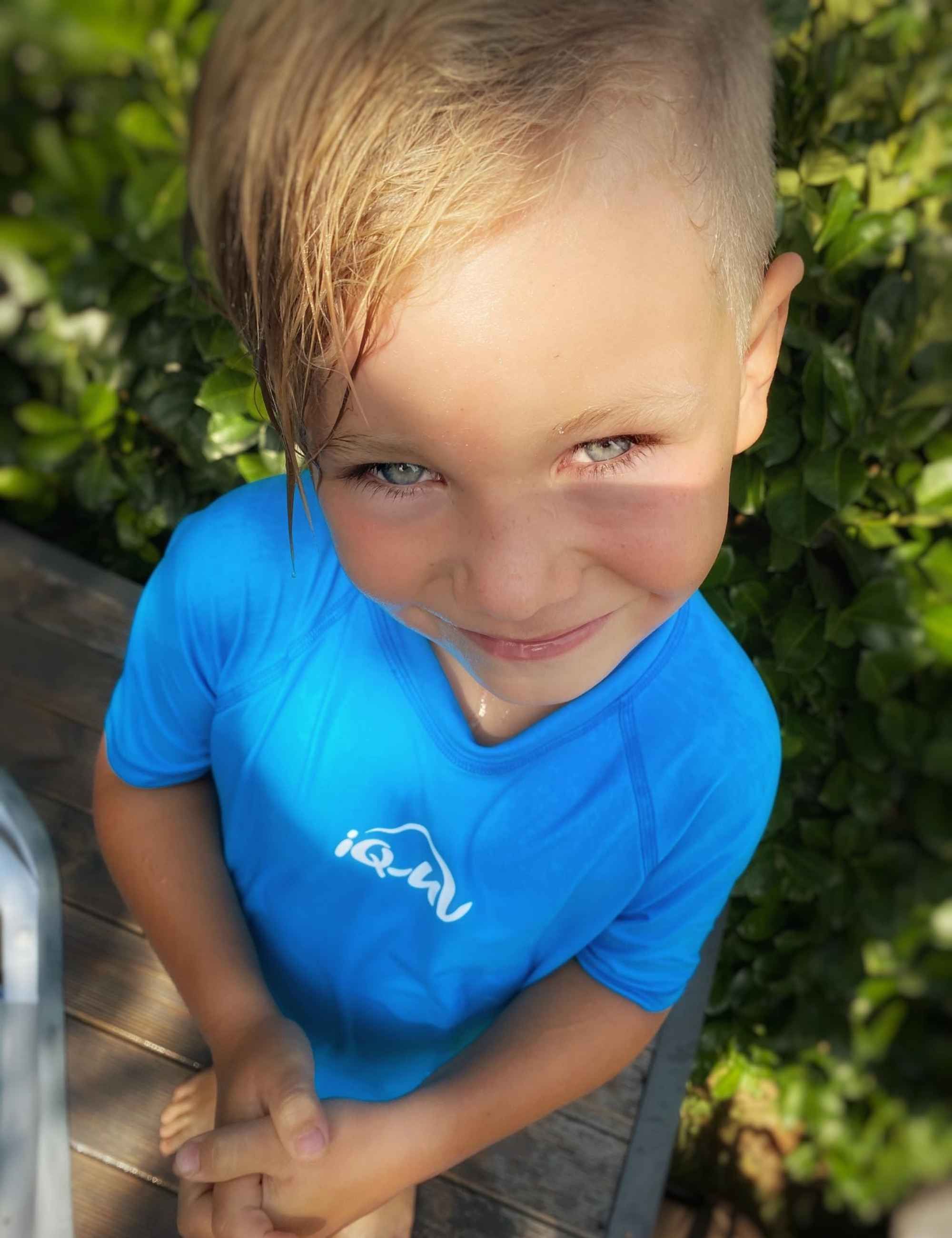 Kind mit blauem Kurzarm-UV-Shirt. Weißes iQ-UV-Logo. Kind blickt zur Kamera. Hintergrund: grüne Pflanzen.
