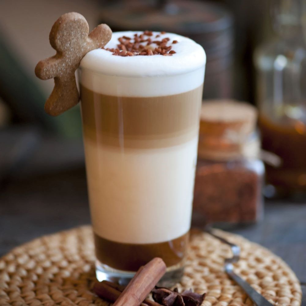 Glas mit geschichteten Getränken: Milch, Kaffee, Sahne. Lebkuchenmann-Keks. Zimtstangen und Sternanis am Boden.