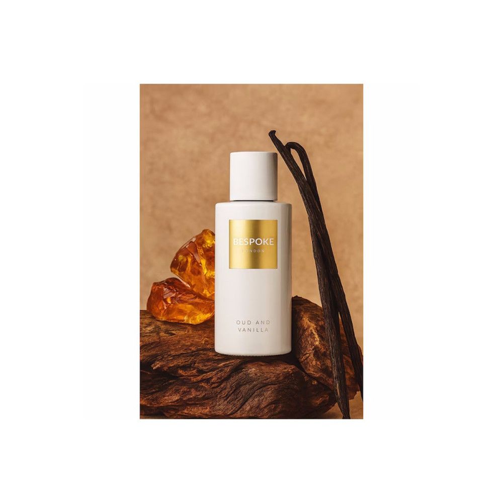 Weißes Parfumflakon mit goldfarbenem Etikett. Text: Bespoke, Oud and Vanilla. Auf Holz und neben Vanilleschoten.