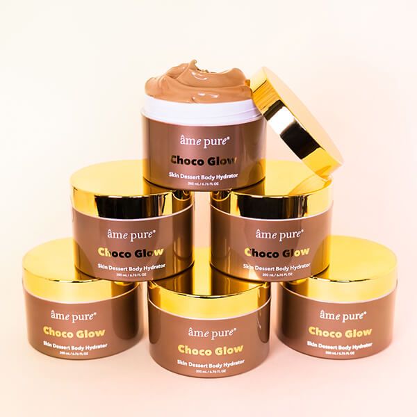 Meerdere bruine potten met gouden deksel. Opschrift: âme pure Choco Glow Skin Dessert Body Hydrator. Opgestapeld.