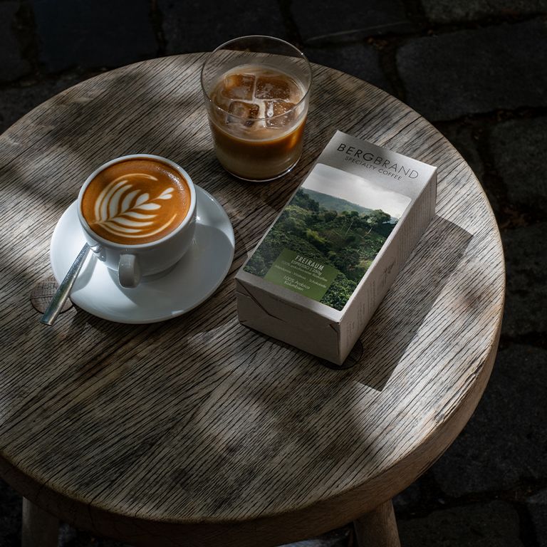 Kaffee mit Latte Art und ein Glas Eiskaffee. Daneben eine Kaffeepackung. Auf einem Holztisch.