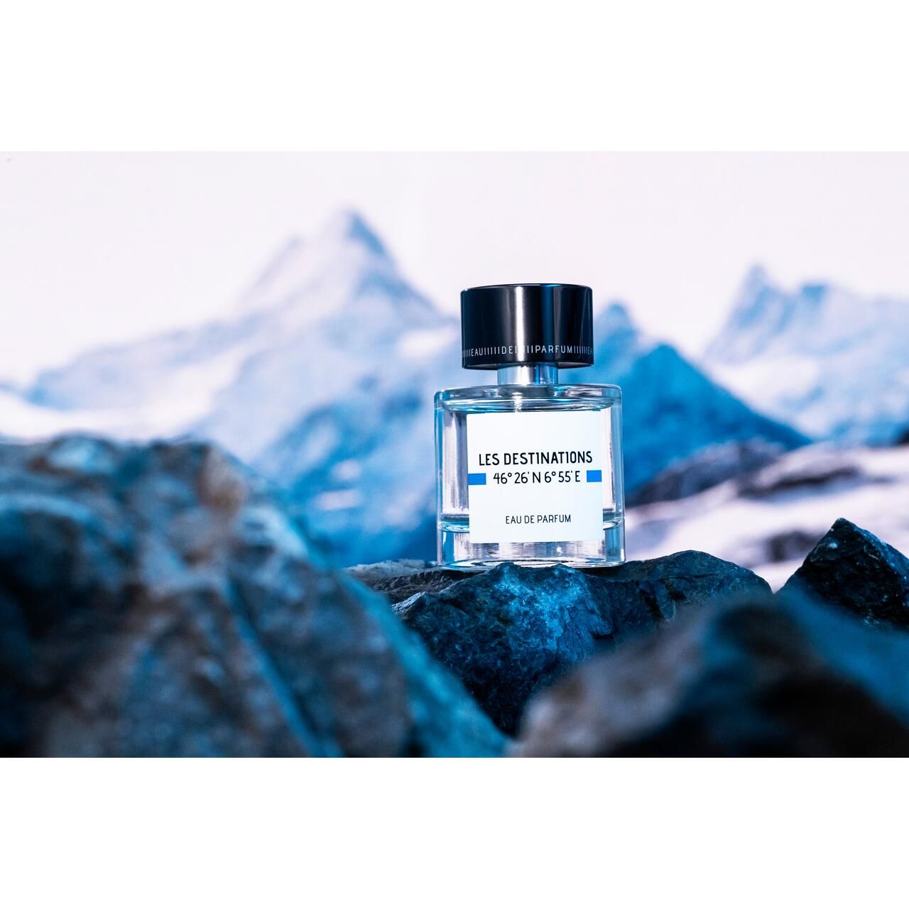 Glasflakon mit schwarzem Deckel vor Berglandschaft. Aufschrift: Les Destinations, 46°26'N 6°55'E, Eau de Parfum.