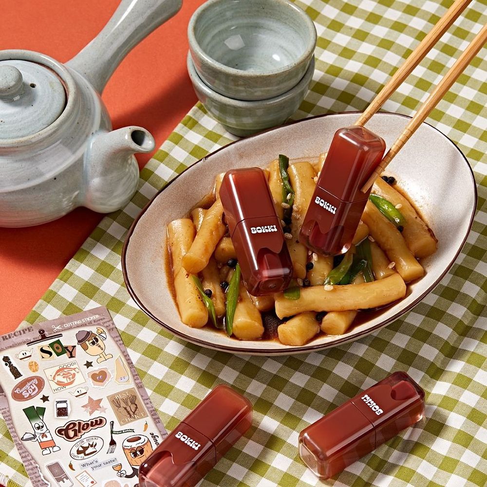 Drei Lippenfarben auf einem Tisch mit Essen. Die Schrift "BOKKI" ist sichtbar. Essstäbchen und Aufkleber sind ebenfalls vorhanden.