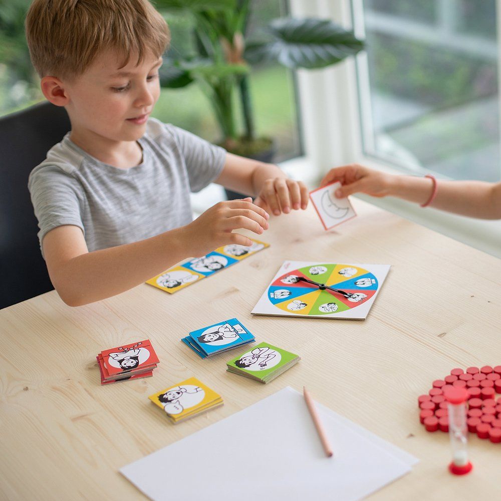 Zwei Kinder spielen am Tisch. Karten, Spielscheibe, Holzplättchen, Stift und Sanduhr sind sichtbar.