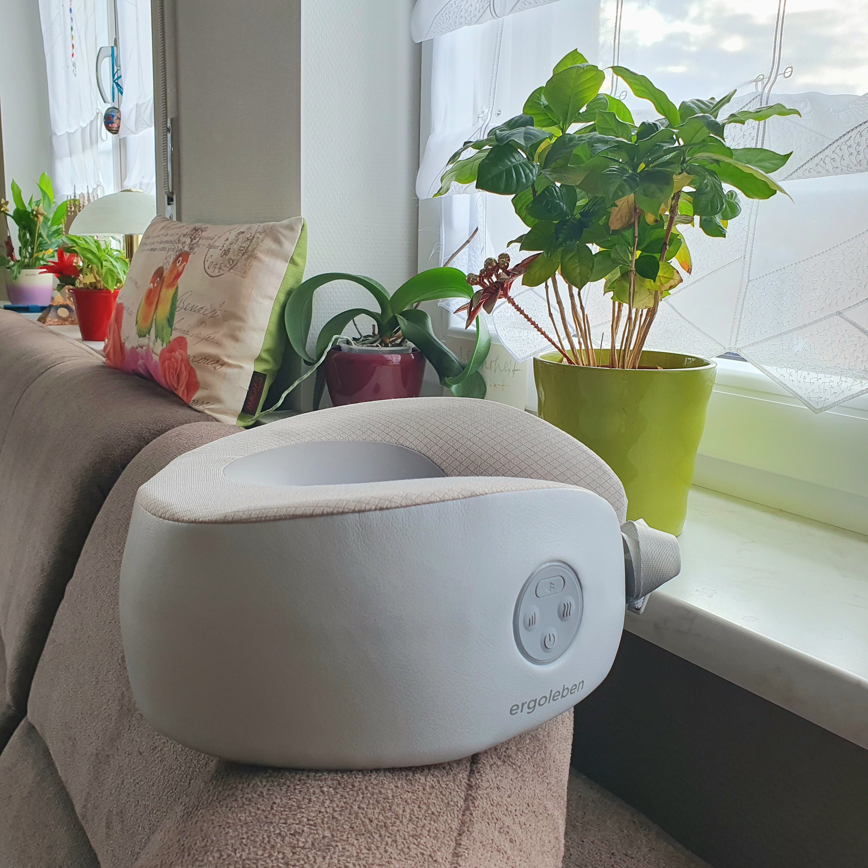 Graues Nackenmassagekissen auf einer Couch. Bedienfeld sichtbar. Umgebung: Fenster, Pflanzen.