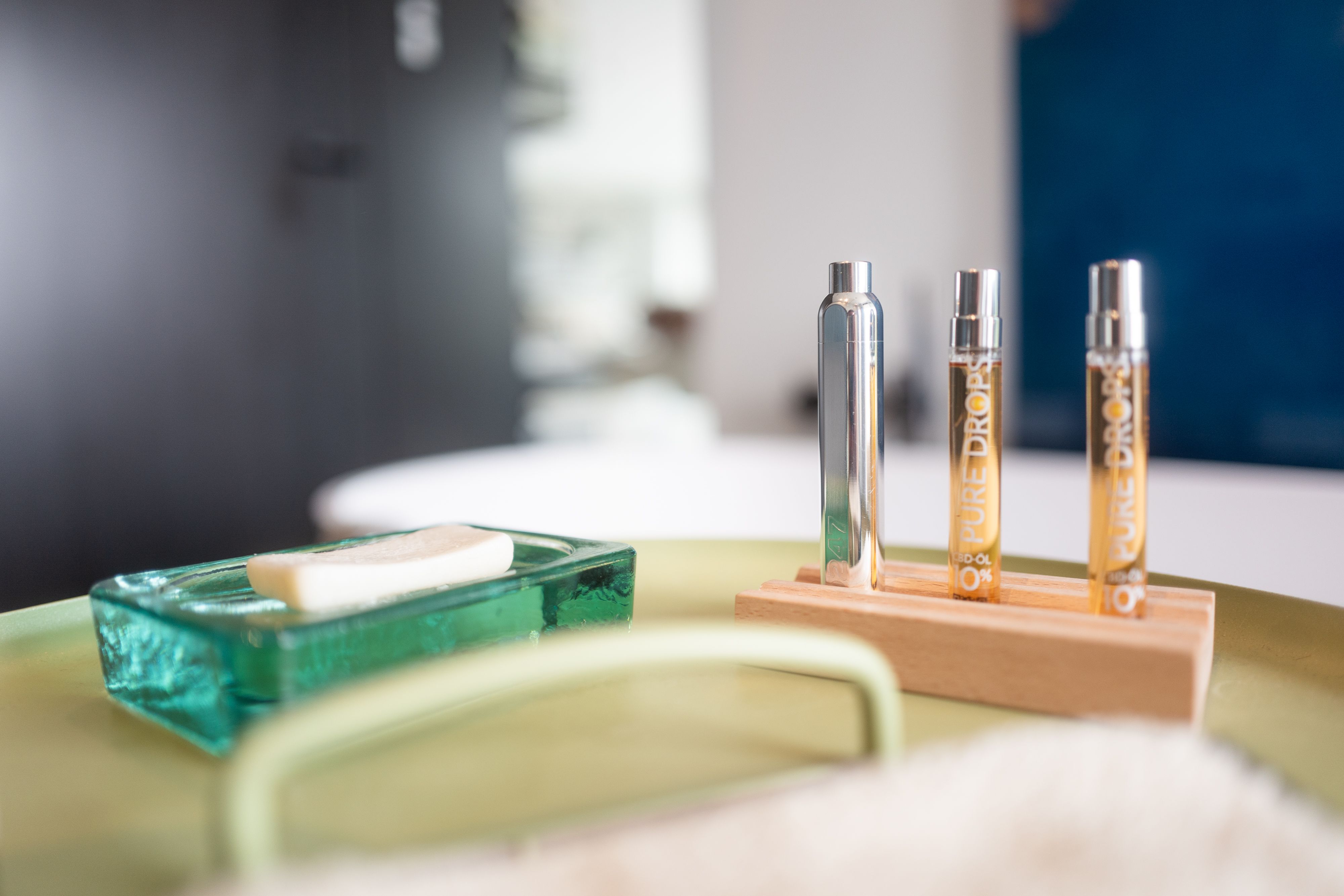 Due flaconi con liquido dorato, un flacone argento, su supporto in legno. Scritta: "PURE DROPS 10% CBD-ÖL".