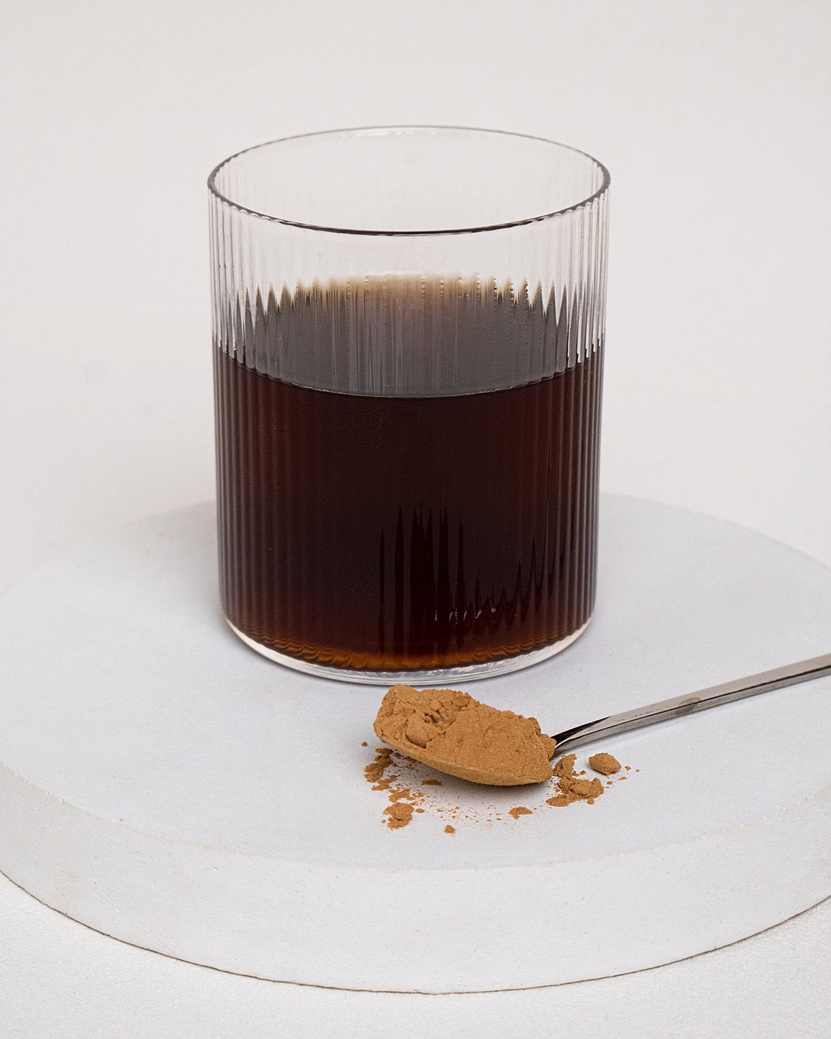 Glas mit dunkler Flüssigkeit, daneben Löffel mit braunem Pulver. Glas steht auf weißer Unterlage. Pulver auf dem Löffel.