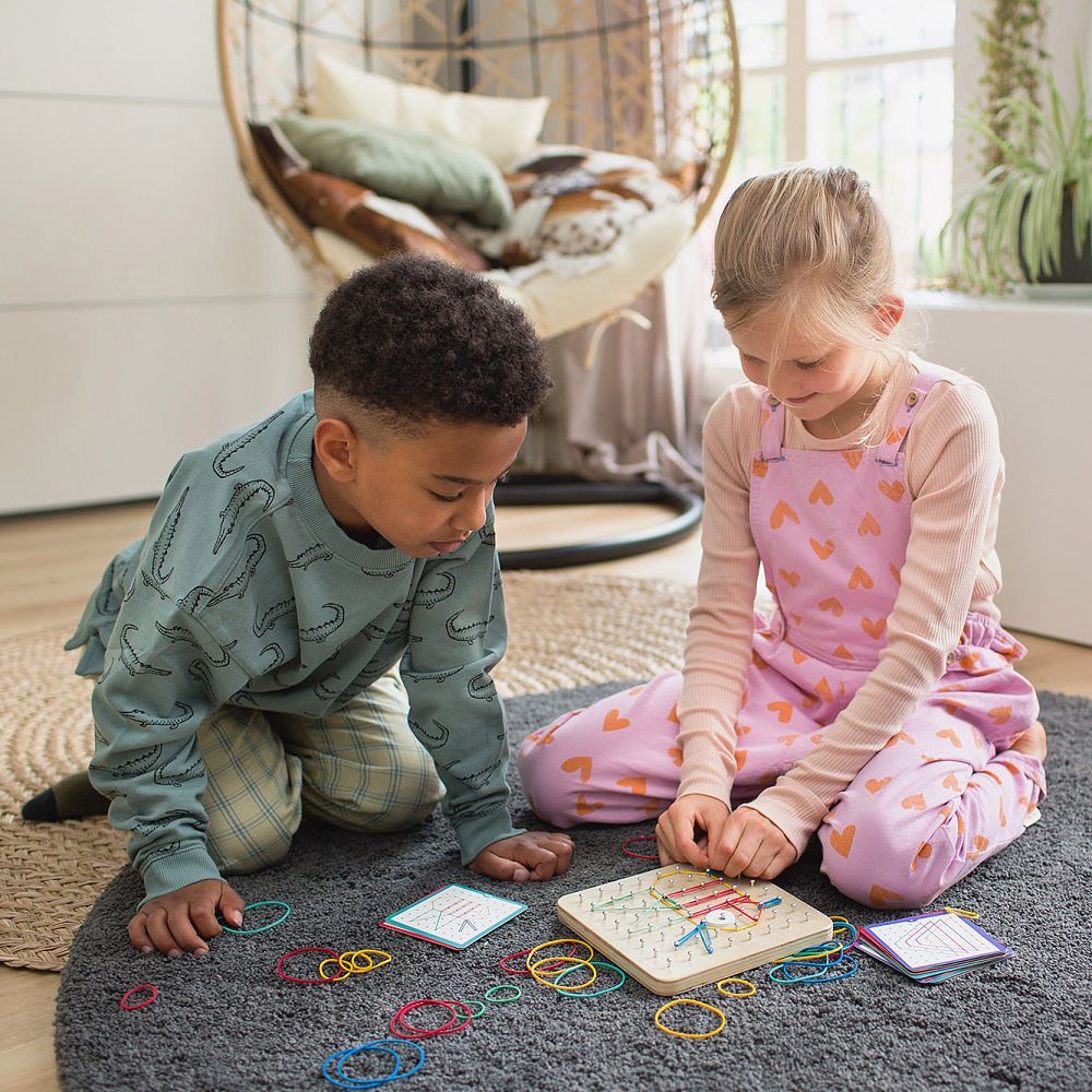 Zwei Kinder spielen mit einem Geoboard. Holzbrett mit Gummibändern. Karten und Gummibänder liegen daneben.