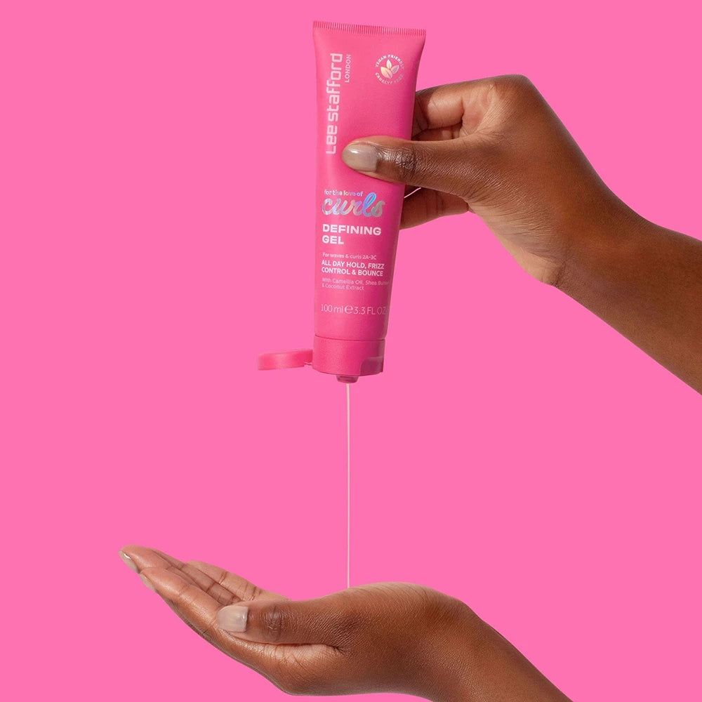 Eine Hand drückt LEE STAFFORD For The Love Of Curls Defining Gel aus einer rosafarbenen Tube in die Handfläche.