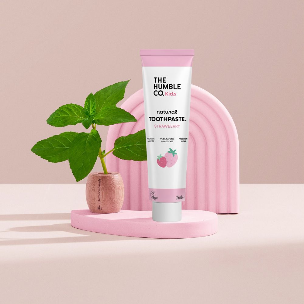 Zahncreme-Tube für Kinder auf rosa Podest. Rosa Hintergrund mit Minze. Produkt mit Logo und Schriftzug "Kids Zahnpasta Erdbeere".