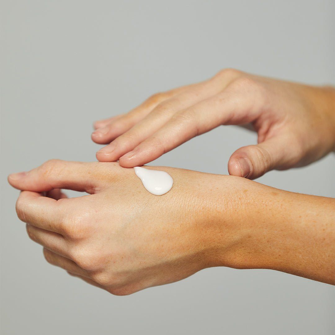 Mani che applicano una piccola quantità di crema bianca sul palmo. La crema viene delicatamente massaggiata. La pelle è chiara e la scena è ripresa su uno sfondo neutro.