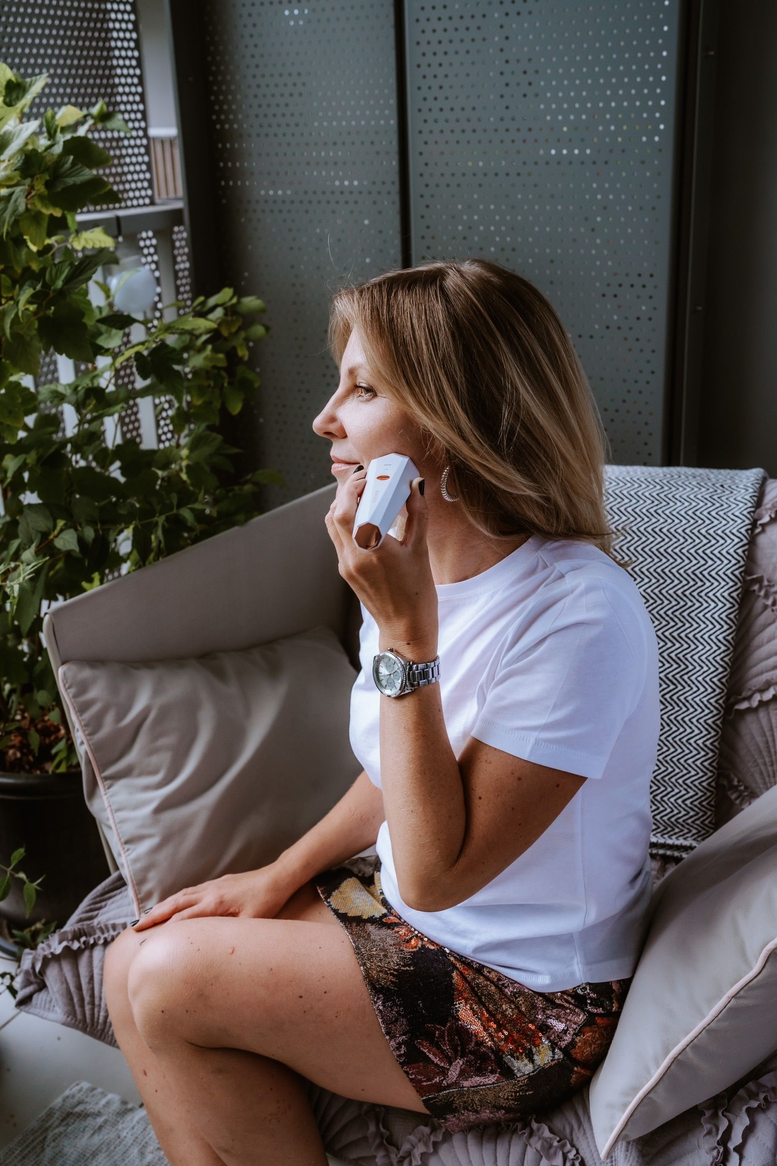 Frau hält ein weißes Gerät an ihr Gesicht. Sie sitzt auf einem Sofa im Freien. Sie trägt ein weißes T-Shirt.