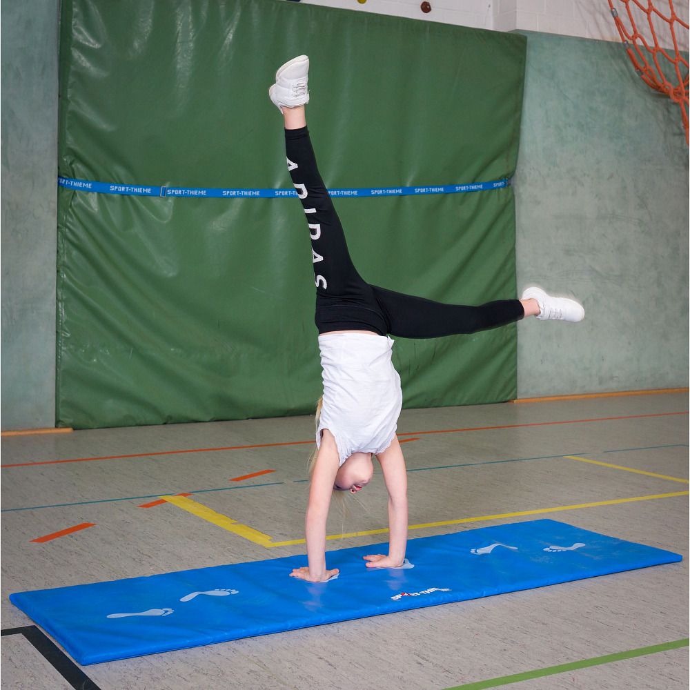 Kind macht Handstand auf blauer Matte. Turnhalle im Hintergrund.