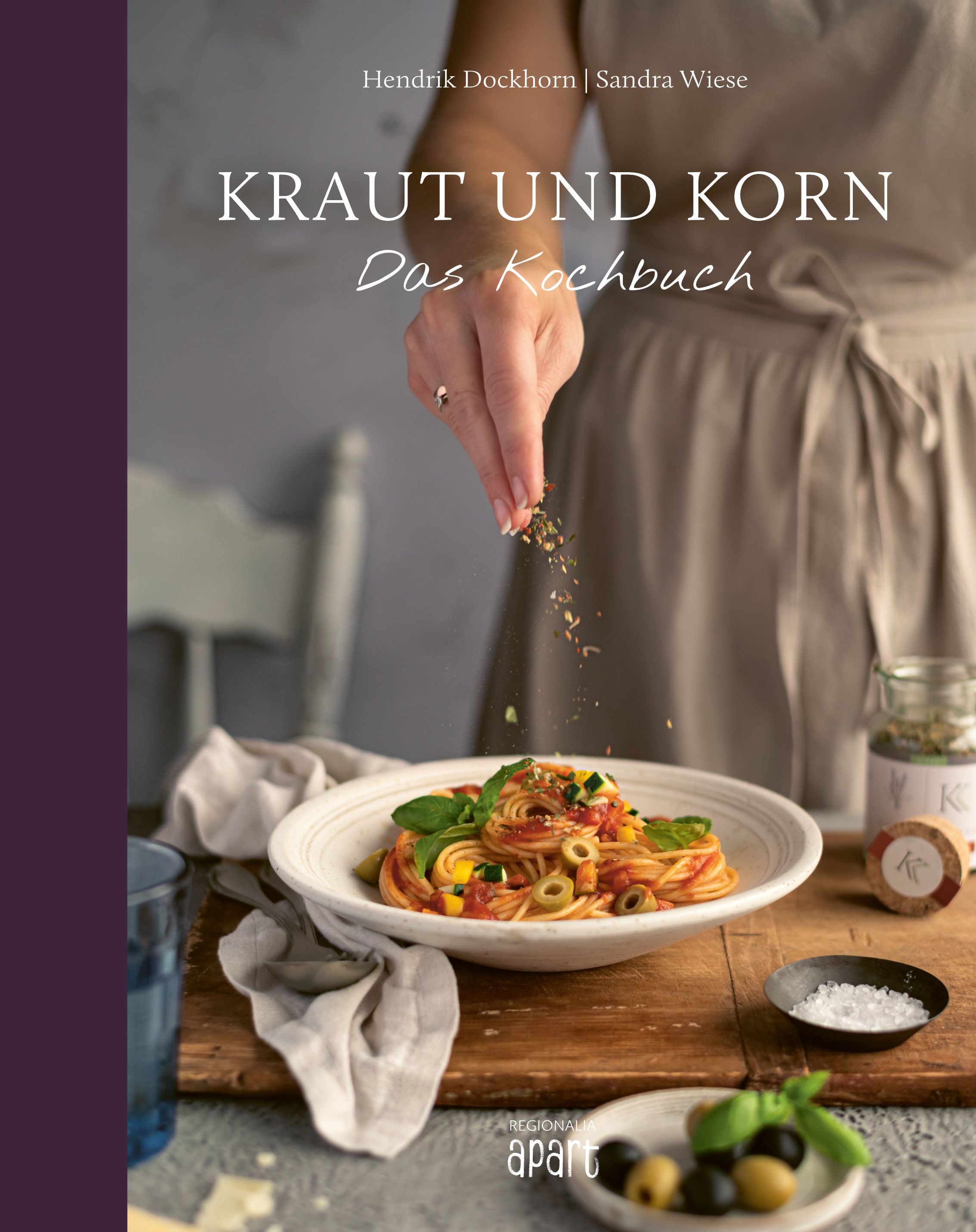Buchcover mit Titel "Kraut und Korn". Eine Hand streut Gewürze über ein Gericht. Auf dem Tisch: Pasta, Oliven, Salz und Kräuter.