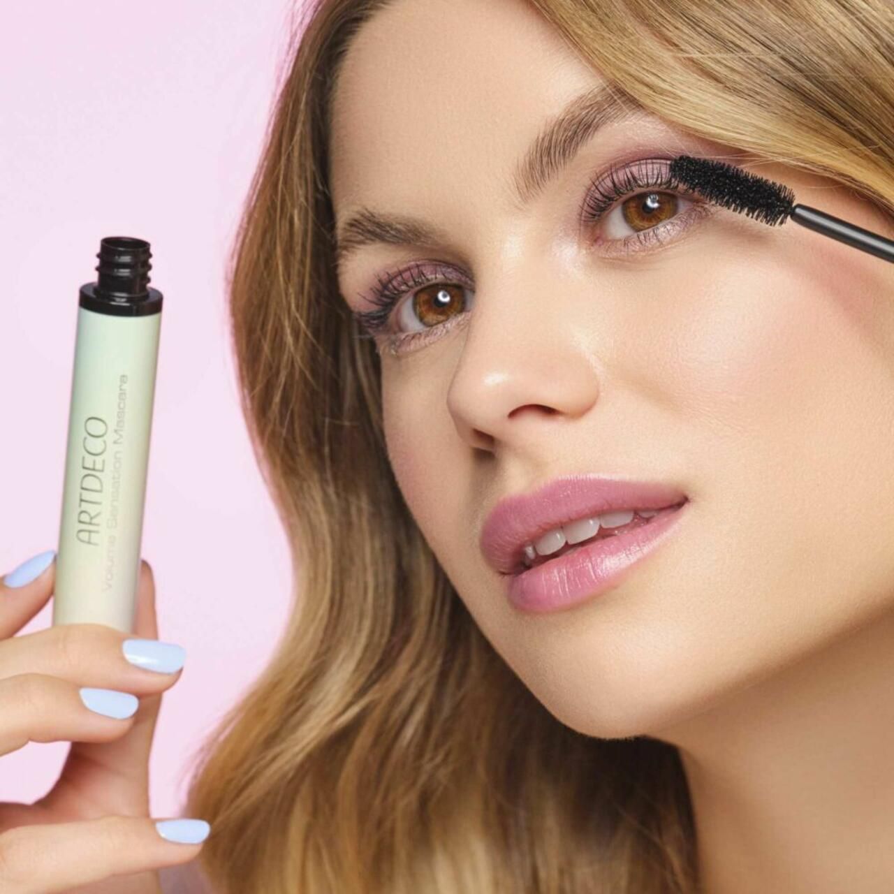 Frau trägt Mascara auf. Artdeco Volume Sensation Mascara. Tube in der Hand. Aufschrift: Artdeco, Volume Sensation Mascara.