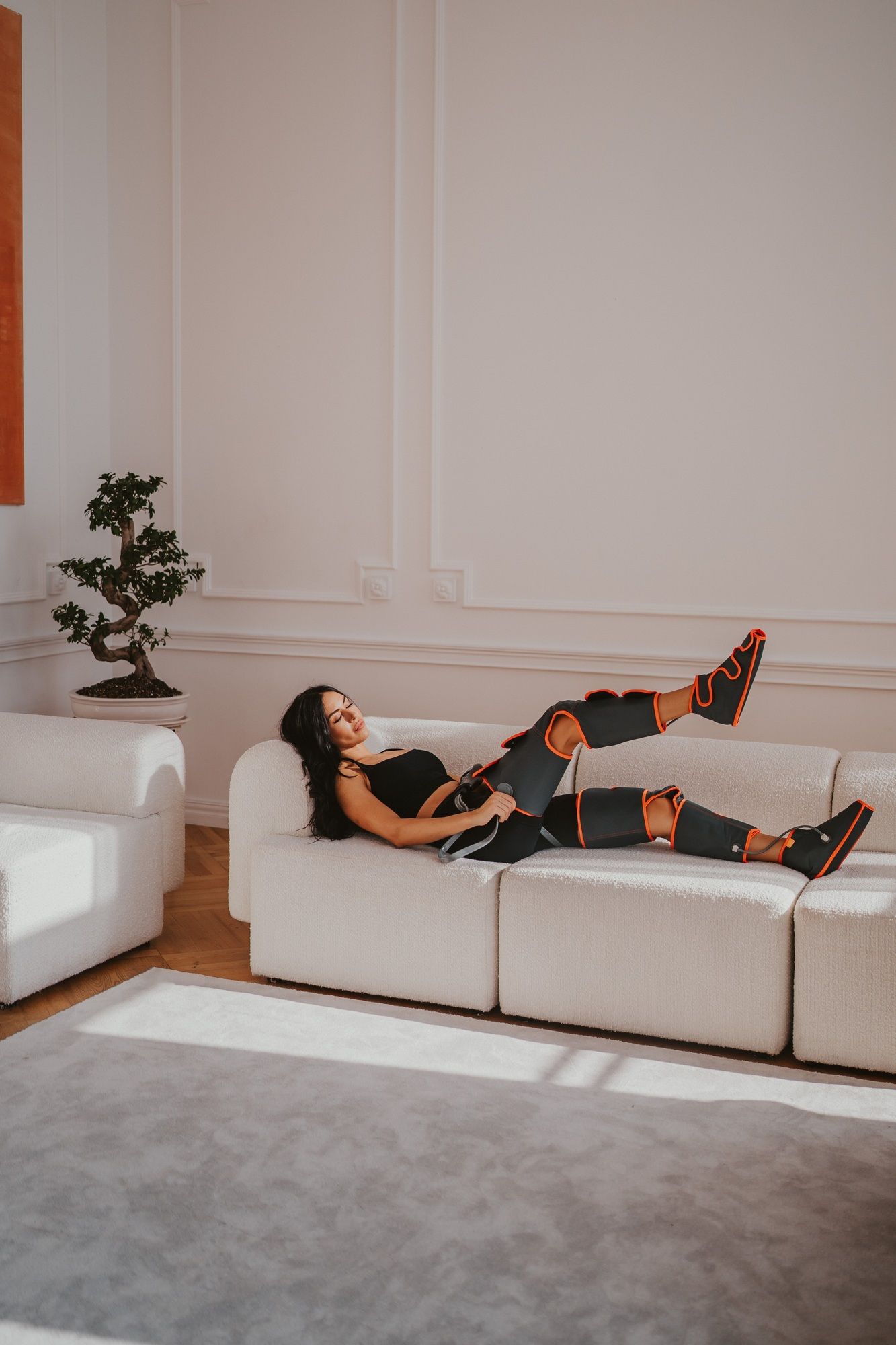 Frau auf Sofa mit Beinmassagegerät. Gerät grau mit orangefarbenen Akzenten. Frau trägt schwarze Kleidung. Wohnzimmer-Umgebung.