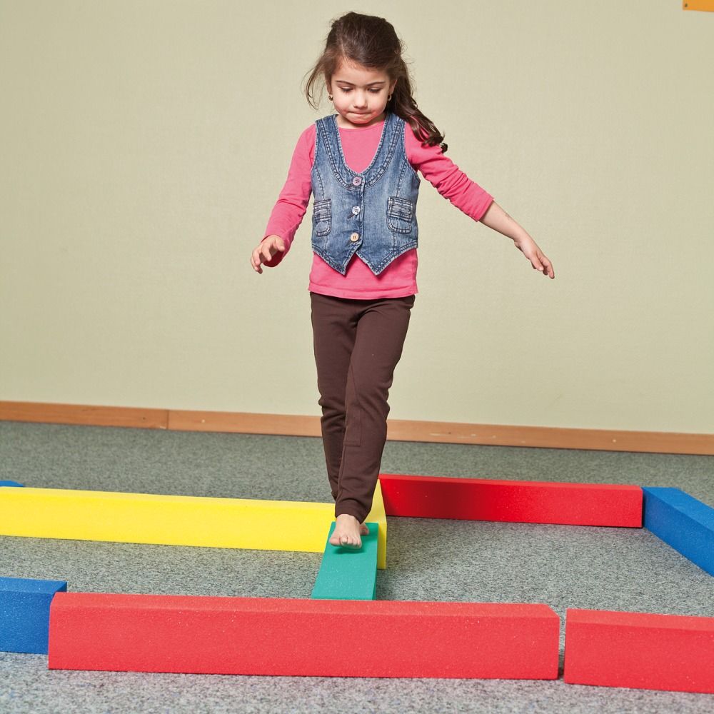 Kind balanciert barfuß auf farbigen Schaumstoffquadern. Gelb, rot, blau und grün. Auf dem Boden angeordnet.