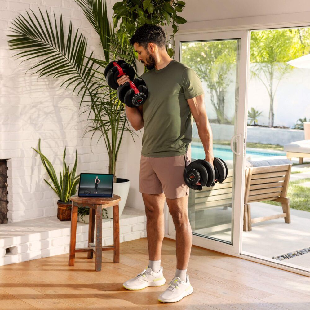 Mann trainiert mit zwei Hanteln. Tablet mit Trainingsvideo. Innenraum mit Möbeln und Pflanzen.