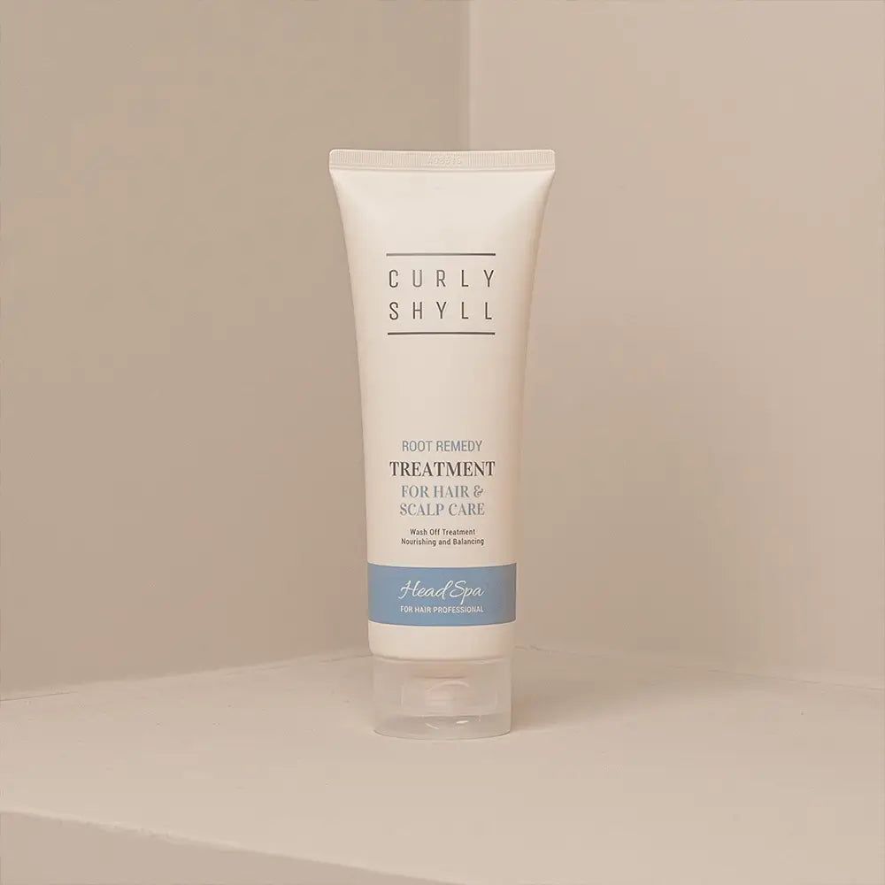 Beige Tube mit Produkttext. Aufschrift: CURLY SHYLL, Root Remedy Treatment, für Haar & Kopfhautpflege. Blaue Leiste: Head Spa. Steht in Ecke.