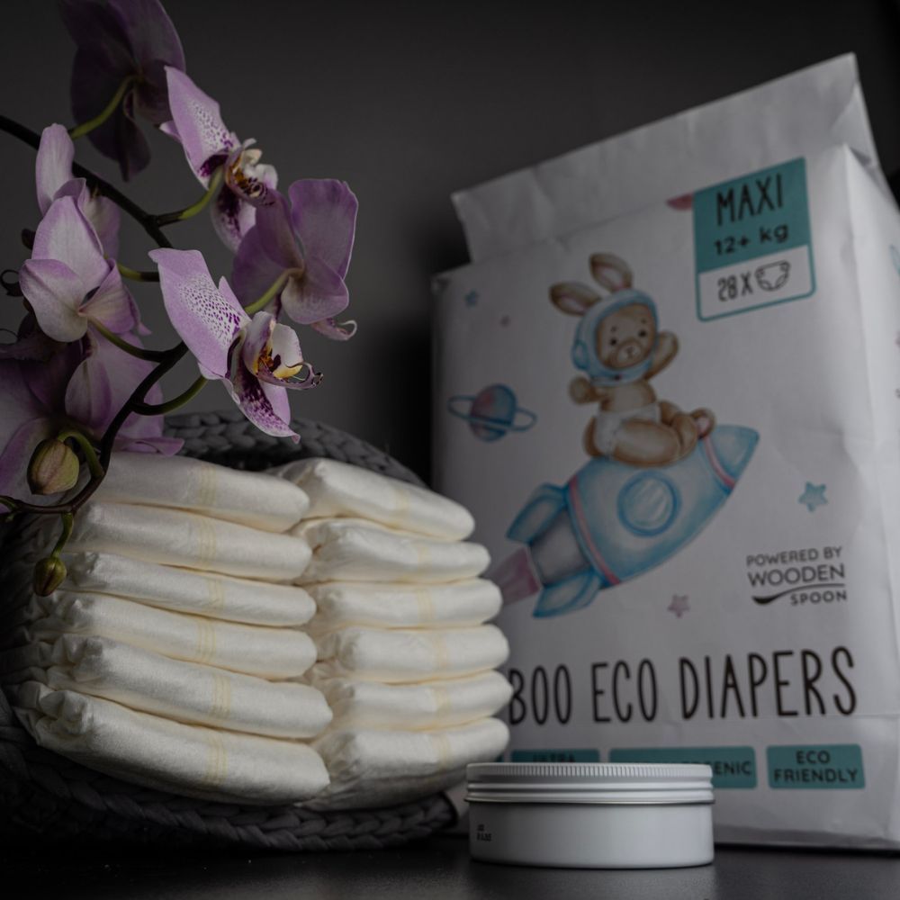 Babywindel-Packung und Stapel Windeln. Aufdruck: Hase auf Rakete, Schriftzug "MAXI 12+ kg", "BAMBOO ECO DIAPERS", "Woopies".