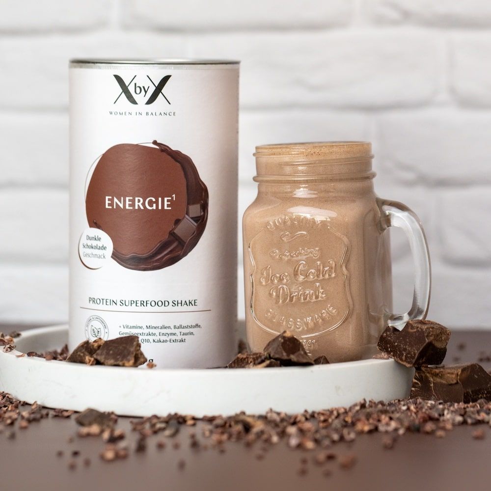 Dose XbyX® Energie Dunkle Schokolade, Glas mit Shake, Schokostücke. Auf Tablett mit Kakaobohnen. Produktname und Logo sichtbar.
