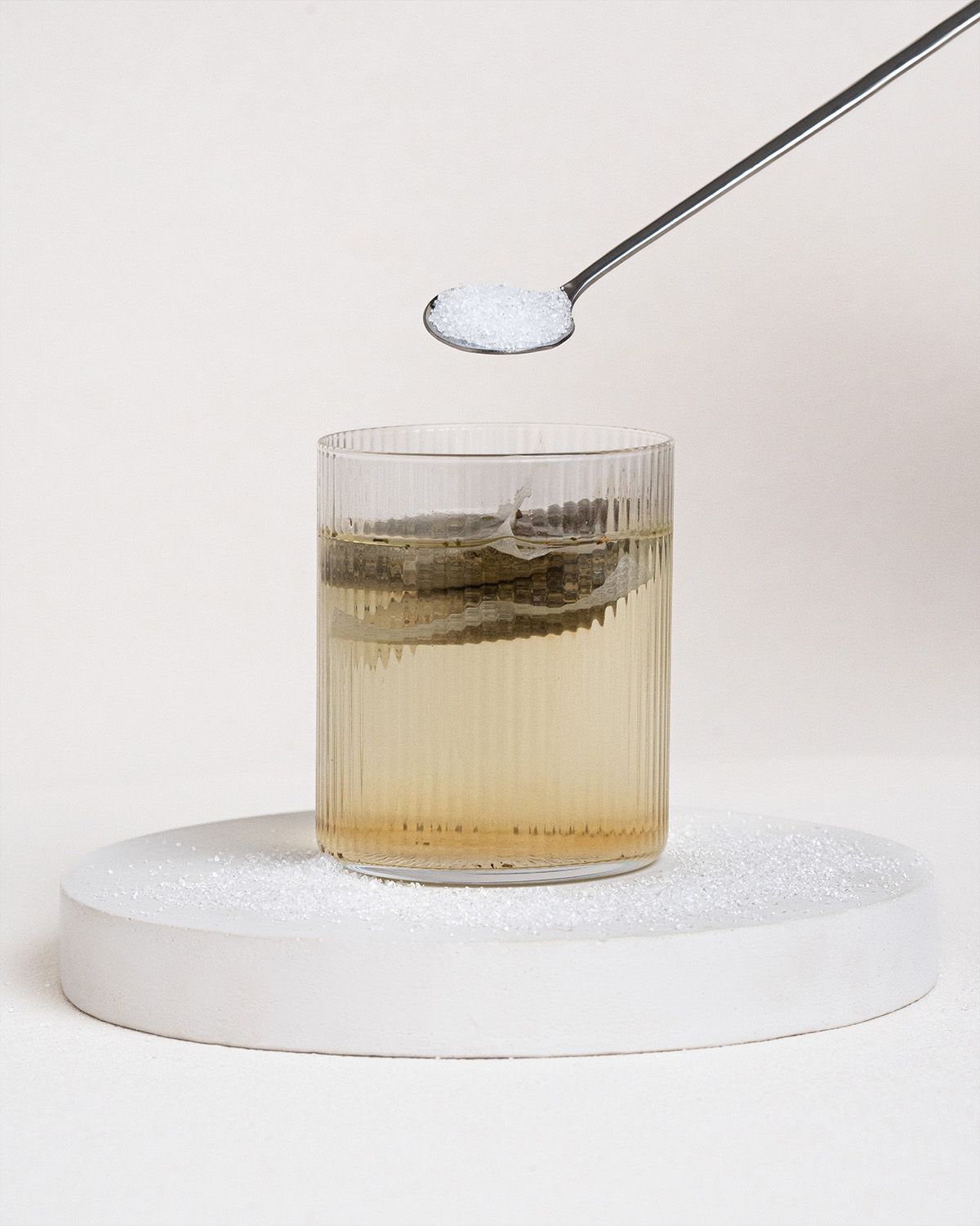Glas mit Flüssigkeit und Teebeutel. Löffel mit Xylit über dem Glas. Auf weißem Podest mit Xylit.