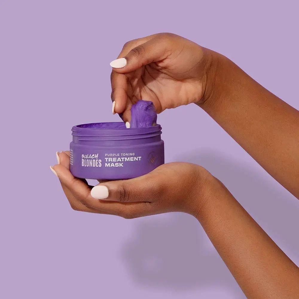 Hände halten einen lila Tiegel mit der Aufschrift Bleach Blondes, Purple Toning Treatment Mask. Produkt wird entnommen.
