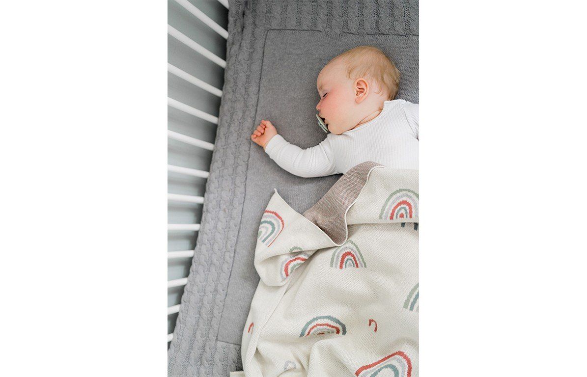 Ein Baby liegt in einem Bett und ist mit einer Decke mit Regenbogenmuster zugedeckt. Beige mit bunten Regenbogenmotiven.
