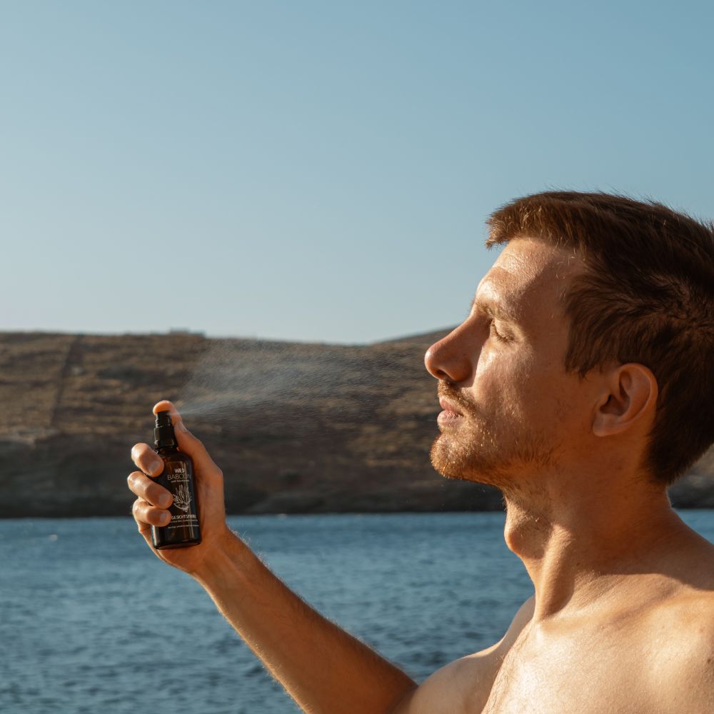Mann sprüht Gesichtstoner. Braune Flasche mit schwarzem Sprühkopf. Text: Wild Baboon. Meer im Hintergrund.