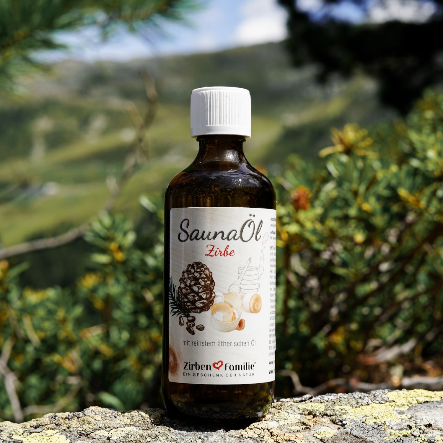 Braune Glasflasche mit weißem Deckel. Etikett SaunaÖl Zirbe. Auf Stein vor grünem Hintergrund.