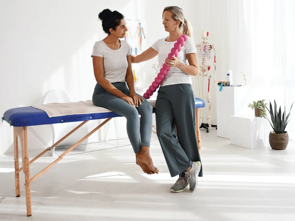 Frau hält pinkfarbenen SPINEFITTER®. Zwei Frauen in einem Behandlungsraum. Eine sitzt auf einer Liege.