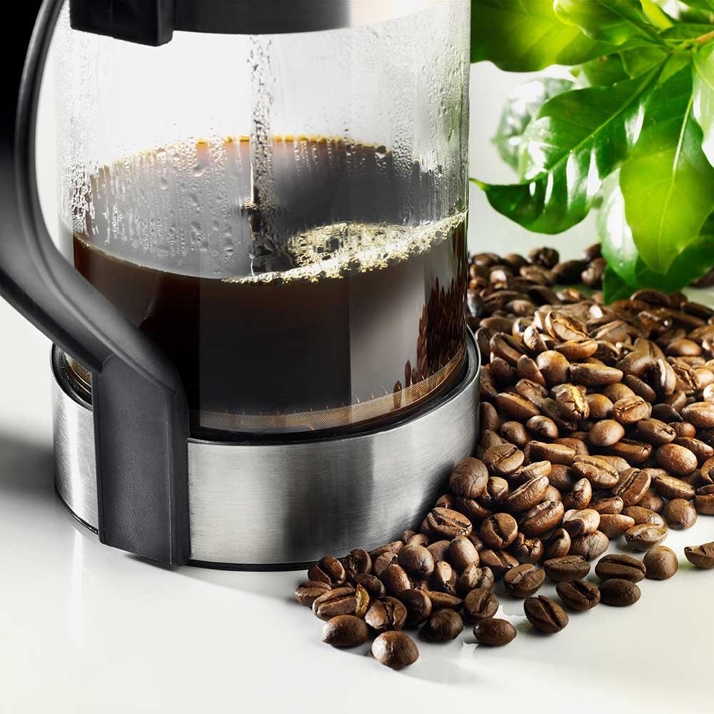 Kaffeebohnen neben Glaskanne mit Kaffee. Blätter im Hintergrund. Edelstahl- und Kunststoffelemente.