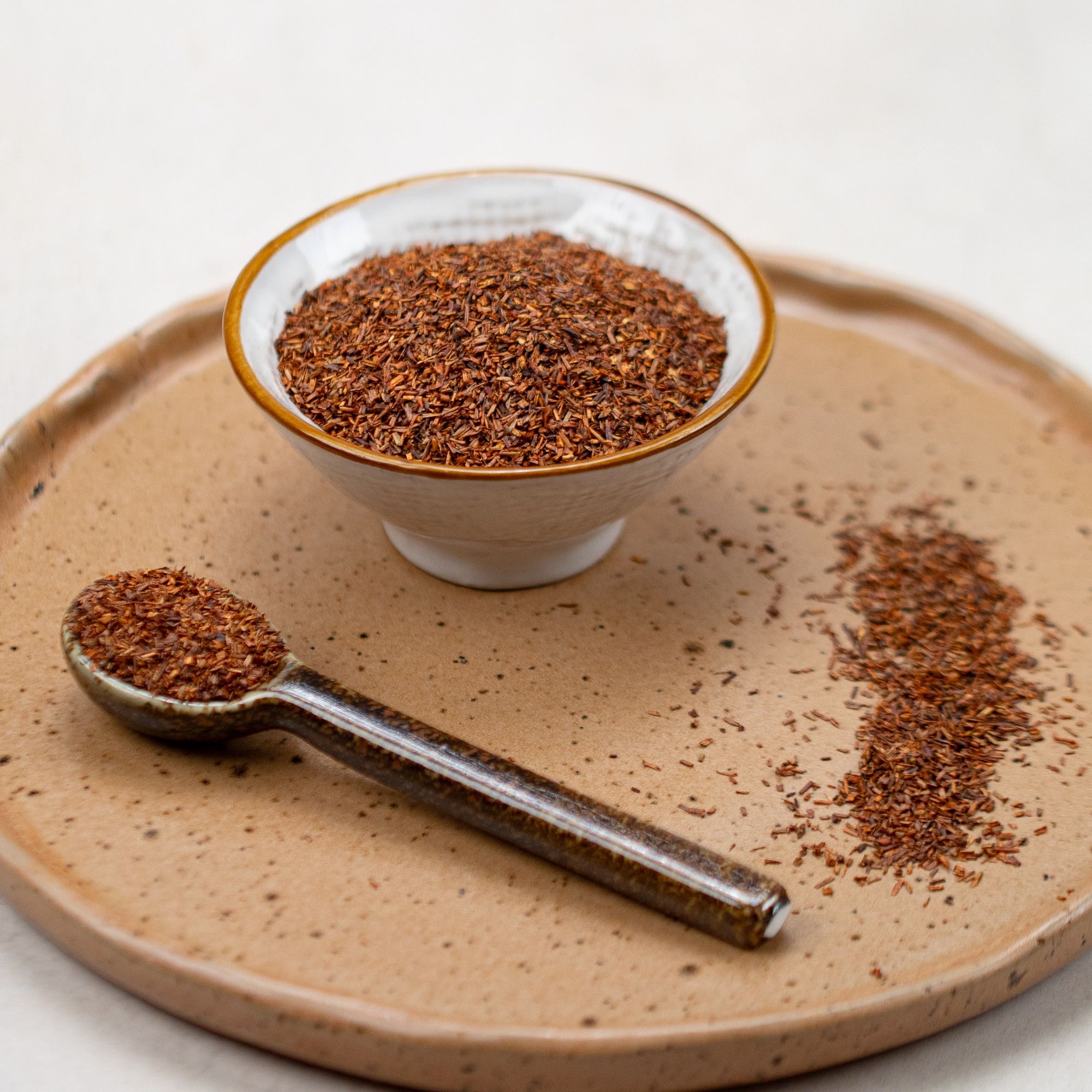 Schale und Löffel mit losem Rooibos-Tee auf einem Teller. Tee ist rotbraun. Teller ist beige.