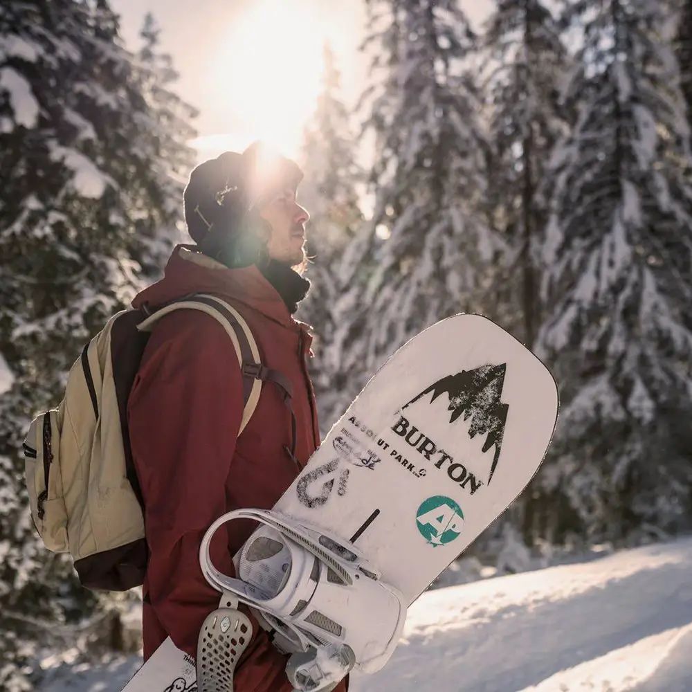 Person mit Rucksack und Snowboard im Schnee. Snowboard mit Burton-Logo und Aufdruck. Winterlandschaft mit Bäumen.