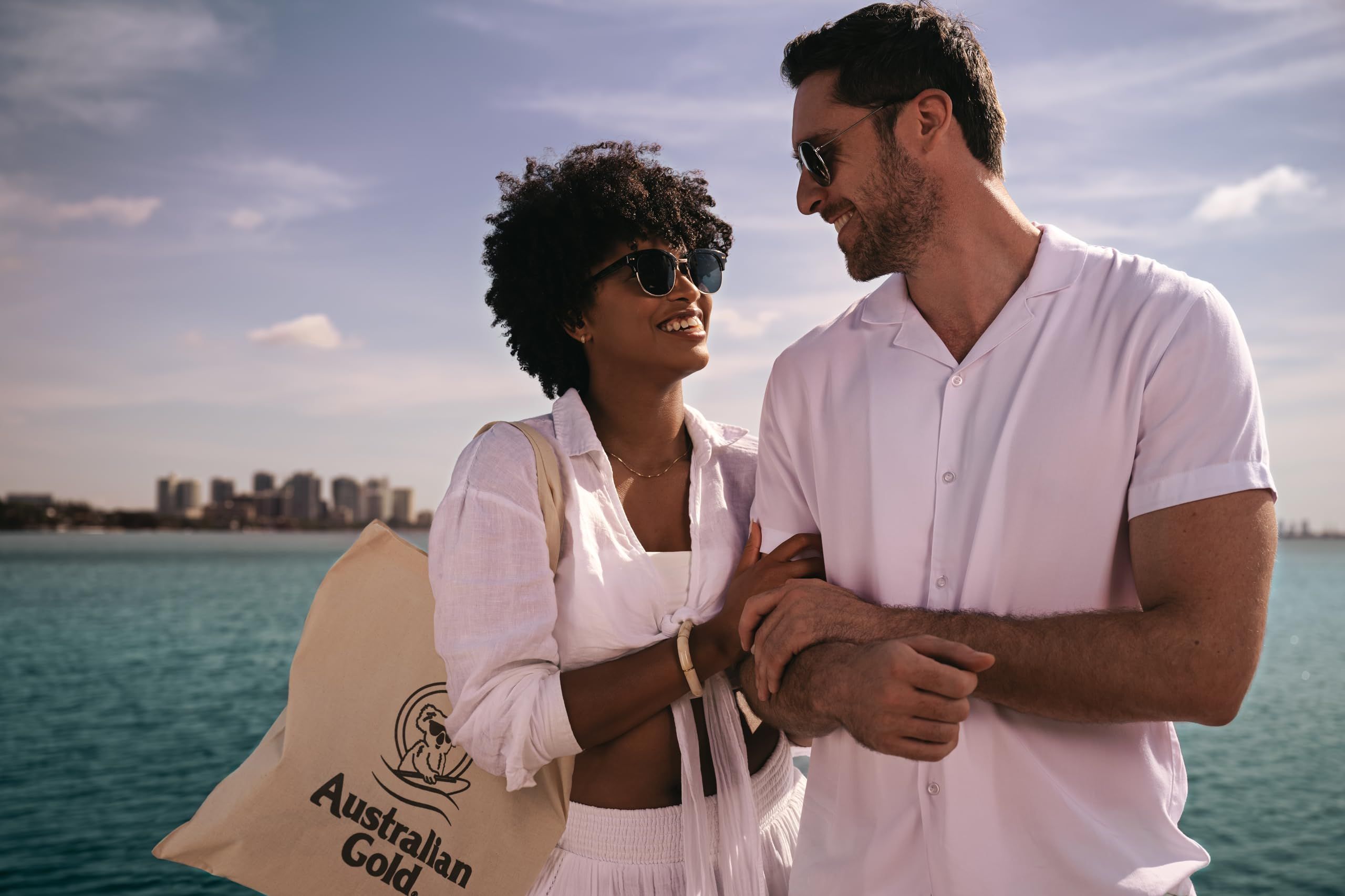 Paar am Meer. Frau mit Sonnenbrille und Mann mit Sonnenbrille. Beide tragen weiße Kleidung. Tasche mit Logo.