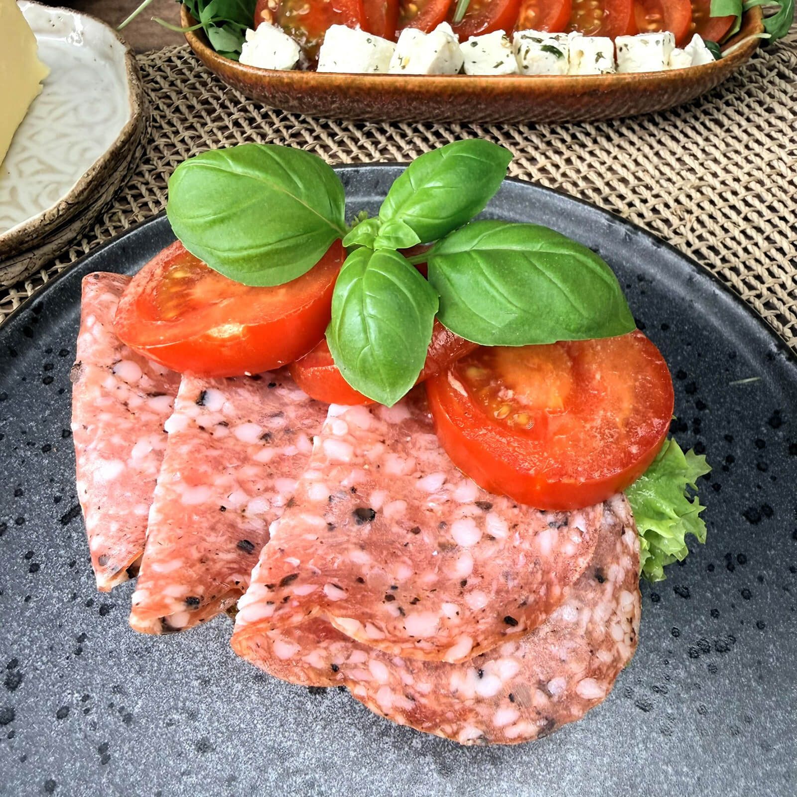 Teller mit Wurstscheiben, Tomaten und Basilikum. Auf dem Teller sind Wurstscheiben, Tomatenhälften und Basilikumblätter angerichtet.