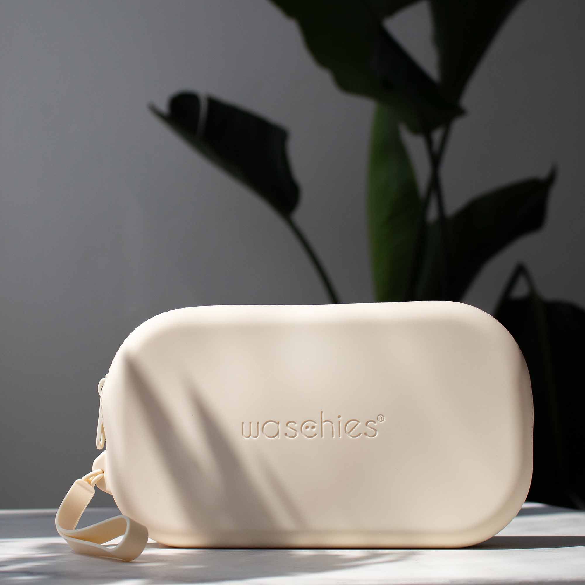 Beige Aufbewahrungstasche mit Reißverschluss und Trageschlaufe. Auf der Tasche steht 'waschies'. Im Hintergrund eine Pflanze.