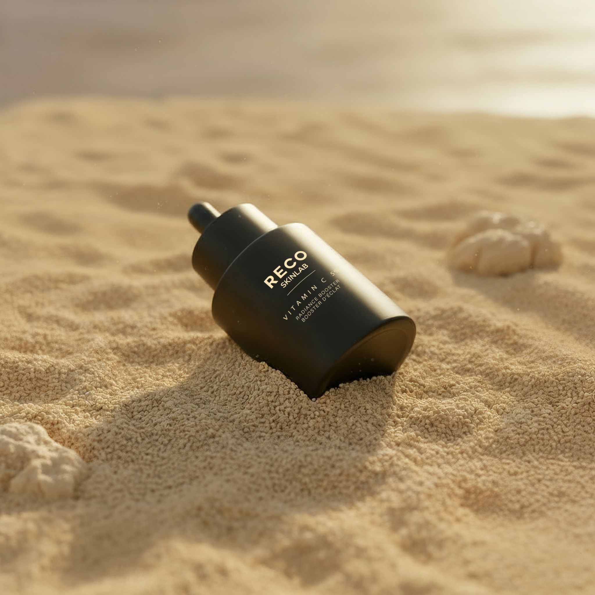Schwarze Flasche mit Pipette, teilweise im Sand. Aufschrift: RECO SKINLAB, Vitamin C, Glow-Booster.