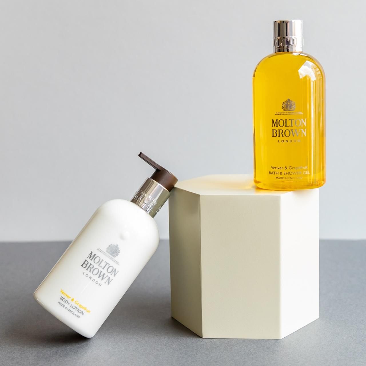 Zwei Molton Brown Produkte auf einem Podest. Eine gelbe Flasche und eine weiße Lotion-Flasche.